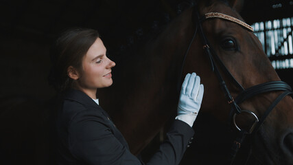 Female horse owner patting her seal brown horse in the stable. Sunlight striking through the...