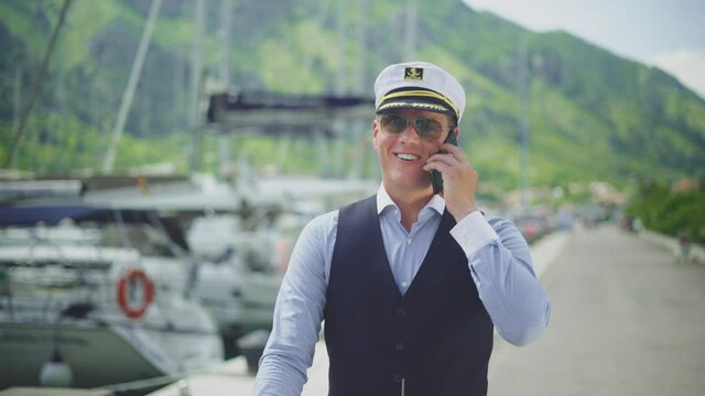 Happy smiling ship captain walking in marina and speaking to cell phone. Summer vacation at sea, sailing and travelling concept.