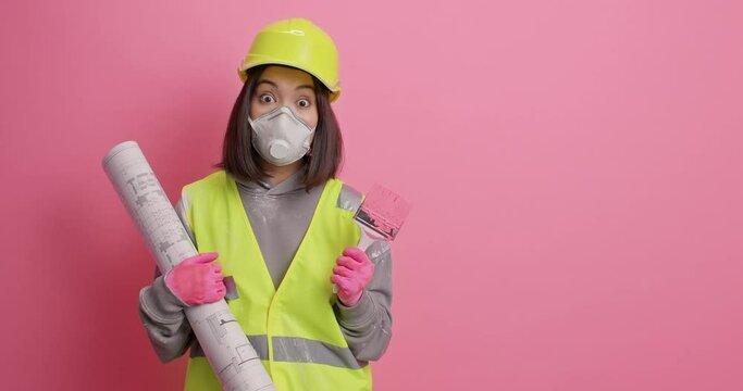 Serious female architect considers future design of room looks attentively hods painting brush and paper blueprint wears protective helmet and uniform poses against pink background blank space