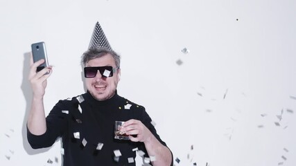 Video greeting. Excited drunk man. Party celebration. Funny guy in sunglasses holiday hat glass drink screaming online congrats on smartphone falling silver confetti isolated white copy space. - Powered by Adobe