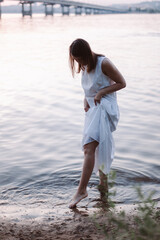 a slender young woman in a white summer dress, lifting the hem of her dress, wets her feet in the water on the sandy beach of the river after sunset.