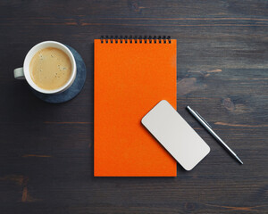 Smartphone, blank orange notepad, coffee cup and pen on wooden background. Responsive design mockup. Top view. Flat lay.