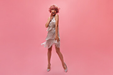 Surprised happy woman in silver shiny dress and high heels poses on isolated. Curly young girl with tattoos on arm looks shocked on pink background.