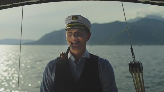 Cheerful sailor captain standing in cockpit and puffing pipe. Mountains in background. Summer vacation at sea, sailing and travelling concept.