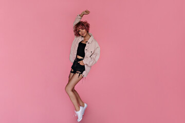 Happy tanned woman in black shorts, cropped top and denim light jacket dances on isolated. Pretty curly girl moves and smiles on pink background.