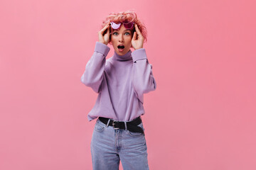 Blue-eyed pink-haired woman in purple sweater and jeans looks surprised into camera on isolated background. Shocked curly girl in denim pants poses on isolated.