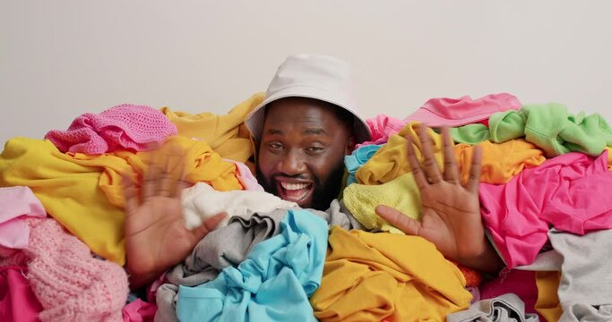 Positive dark skinned guy has cheerful expression sticks head in pile of laundry wears panama wave palms makes like gesture keeps thumbs up brings closet in order sorts clothes poses indoor.