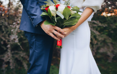 The bride in a white wedding dress and the groom in a blue suit stand against each other and hold a large bouquet of white and pink roses with green foliage.