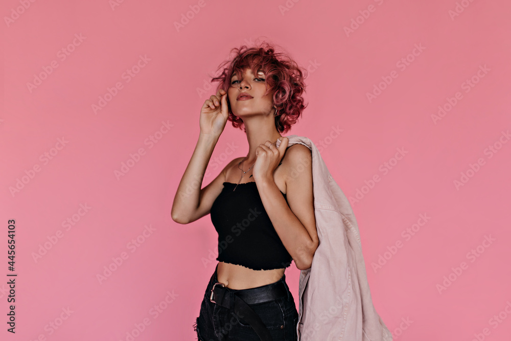 Wall mural Pretty curly girl in stylish black tee and dark shorts looks into camera. Attractive woman holds light pink denim jacket on isolated .