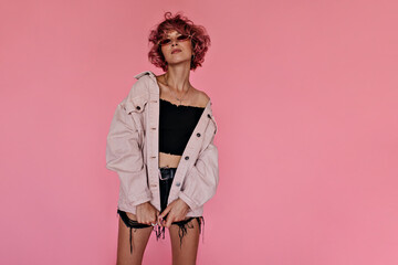 Curly pink-haired woman in dark denim shorts and stylish oversized jacket poses on pink background. Attractive tattooed girl in sunglasses looks gorgeous .