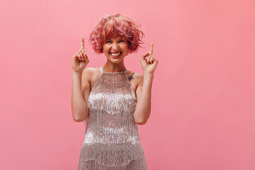 Cheerful curly woman in silver dress smiles widely and points fingers up at place for text on isolated pink background.