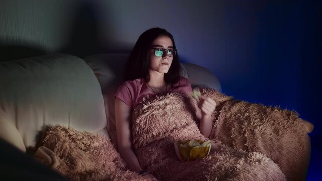 pretty girl watches tv in the evening, switches channels and eats chips, relaxing at home in front of the TV, moving camera, cinematic shot