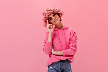 Cheerful pink-haired woman in oversized hoodie and denim pants smiles widely. Young girl in jeans and red sunglasses looks into camera camera on isolated pink background.