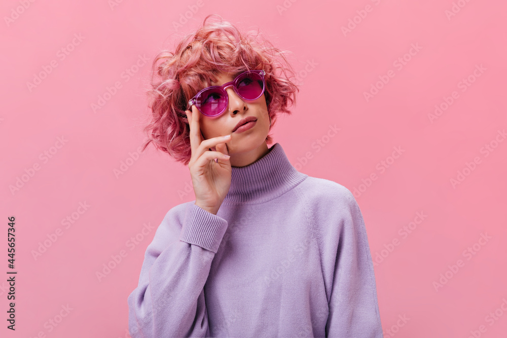 Sticker Portrait of pink-haired curly thoughtful woman in purple sweater and fuchsia sunglasses posing on isolated background .