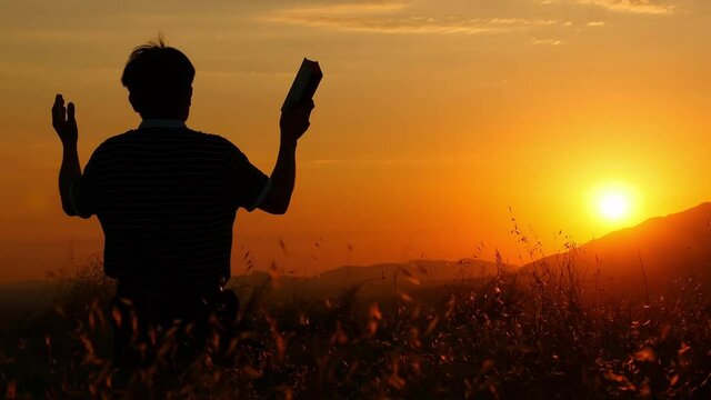 Silhouette of a man praying to God with the Bible in a hand and standing on the grass with arms open at sunset