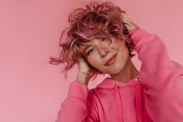 Happy tanned curly woman in bright hoodie ruffles hair. Charming girl with pink hair looks into...