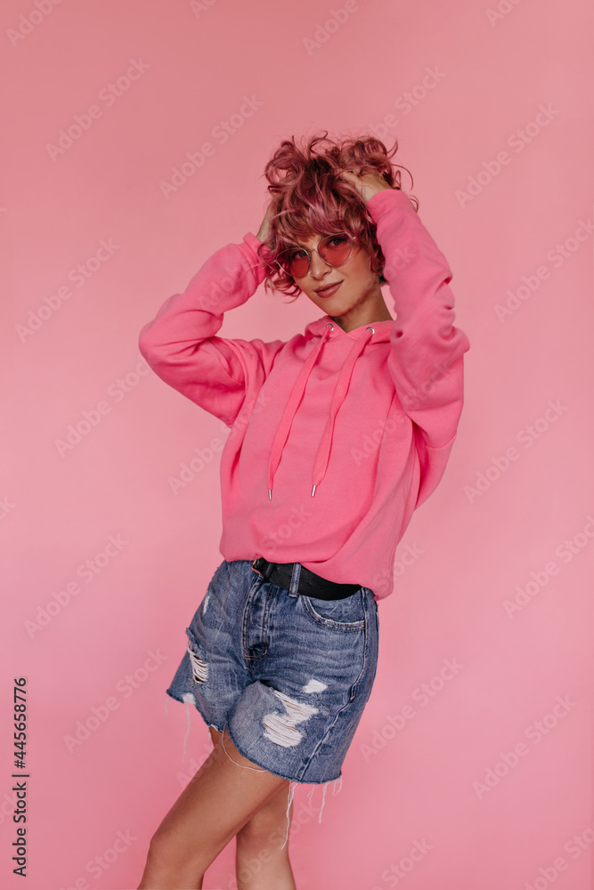 Wall mural Thin tanned girl in hoodie, denim shorts and sunglasses looks into camera on isolated. Curly woman smiles on pink background. - Wall murals