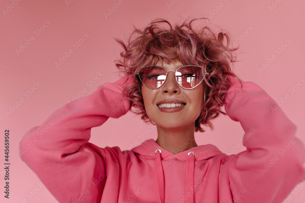 Wall mural Joyful woman in good mood poses on isolated. Portrait of happy young woman in bright sunglasses and pink hoodie smiling .