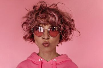 Cool young woman in pink hoodie makes funny face. Charming good-humored girl in sunglasses looks...