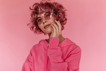 Attractive pink-haired woman in bright colorful sunglasses and fuchsia hoodie looks into camera and touches her face softly on isolated background.