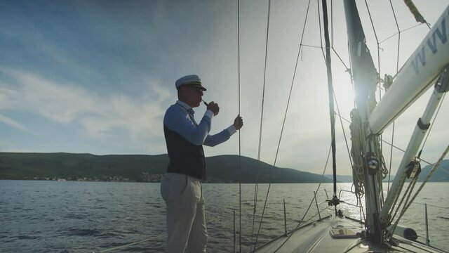 Sailor puffing pipe while cruising calm water at sunset. Standing at deck and holding rope Summer vacation at sea, sailing and travelling concept.
