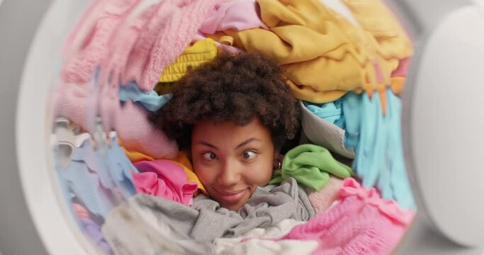 Funny dark skinned curly woman does laundry at home drowned in pile of multicolored clothes in washing machine makes funny grimace crosses eyes smiles broadly busy doing daily domestic routines