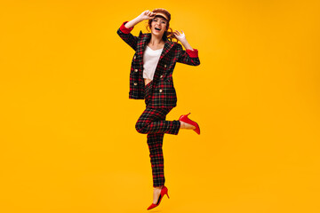 Fototapeta na wymiar Beautiful lady in stylish suit, cap and eyeglasses jumping on orange background. Happy woman in modern outfit and red shoes posing..