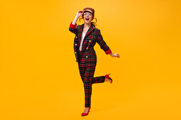 Pretty girl in eyeglasses, cap and suit jumps on orange background. Joyful woman in cap and round eyeglasses posing on isolated backdrop..
