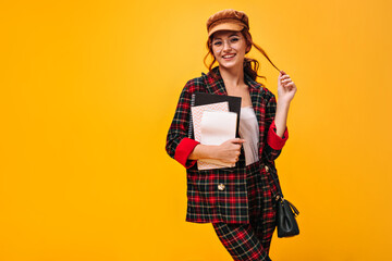 Coquettish woman in plaid outfit poses with notebooks. Attractive girl in brown cap, eyeglasses and modern suit looking into camera..