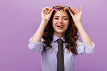 Charming lady in blue shirt taking off eyeglasses and laughing. Pretty woman with wavy hairstyle in black tie posing on purple background..