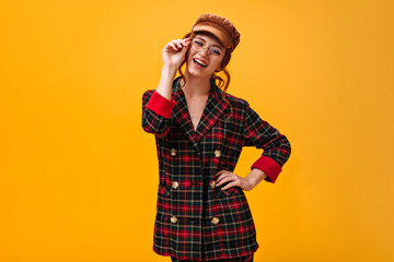 Happy woman in eyeglasses, cap and plaid jacket smiling on orange background. Modern lady with ginger hair in fashionable suit posing..