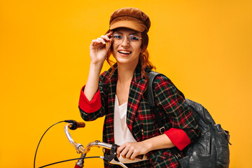 Happy girl in plaid jacket and eyeglasses poses with bicycle on orange background. Joyful woman with red hair in cap and stylish clothes smiling..