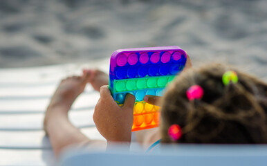 irl 5 years old in a swimsuit lying on a sun lounger by the sea on a sandy beach plays Pop It anti-stress on a hot sunny day.
