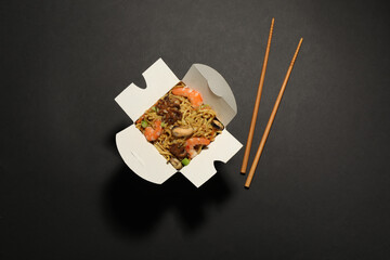 Box of wok noodles with seafood and chopsticks on black background, flat lay