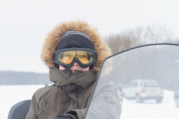 Fototapeta na wymiar Portrait of a teenage boy in yellow safety glasses and a hood with fur in winter on a snowmobile
