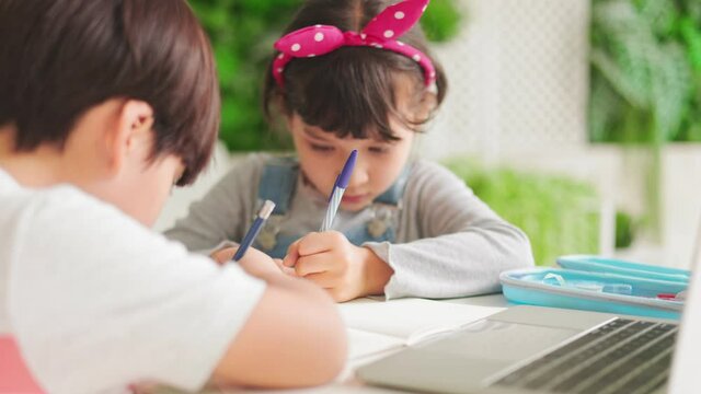 Small mixed race  girl sit at desk study online on laptop, smart little kid handwrite in notebook learning using internet lessons on quarantine, homeschooling concept.
