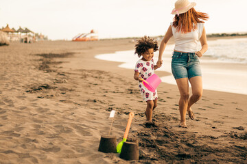 Caucasian mother and african daughter having fun running on a sandy beach. Mum and son building a sandcastle at sea.