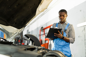 African American auto mechanic man in overalls and holding a notebook in his hands while standing...