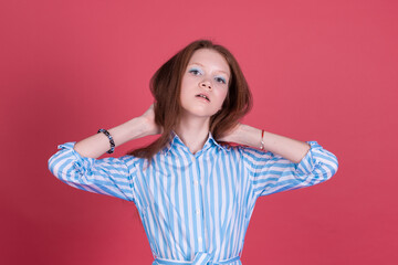 Little kid girl 13 years old in blue dress isolated on pink background serious holding hair look to camera