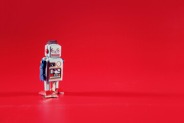 A solitary tin toy robot, standing still over a red backdrop, with copy space on the right.
