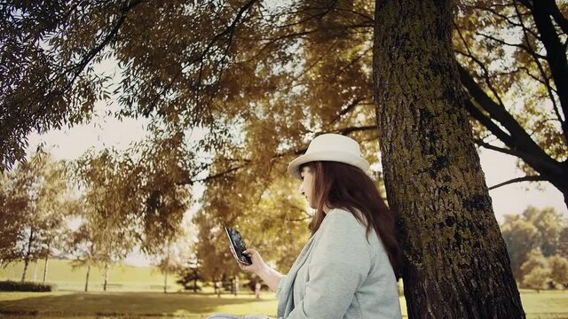 brunette woman in a hat sits in the park in autumn under a tree and uses the phone, read some information, chat with friends, cinematic shot