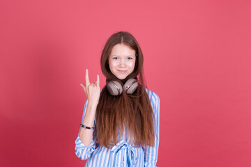 Little kid girl 13 years old in blue dress with brackets isolated on pink background in wireless headphones shows rockstar gesture