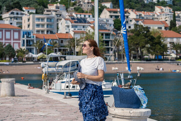Young woman tourist in a hat, sunglasses, blue skirt and white blouse walking on the promenade of a...
