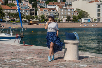 Young woman tourist in a hat, sunglasses, blue skirt and white blouse walking on the promenade of a...