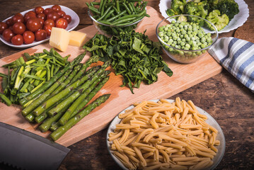 Ingredients for cooking pasta Primavera - pasta in a dish, beans, asparagus, broccoli, peas, tomato and a slice of cheese on a chopping board