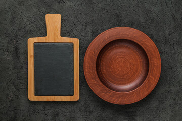 Brown wooden cutting board with slate stone, empty brown clay plate on black stone table. Top view...