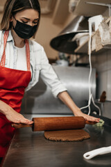 Beautiful young female seller with protective face mask working in a confectionery shop or bakery. She is making delicious sweets. Coronavirus and small business concept.