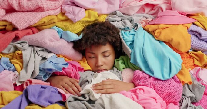 Tired sleepy young curly woman drowned in pile of laundry feels fatigue keeps eyes closed cleans out wardrobe collects clothes for donation sorts clothing does laundering busy with domestic work
