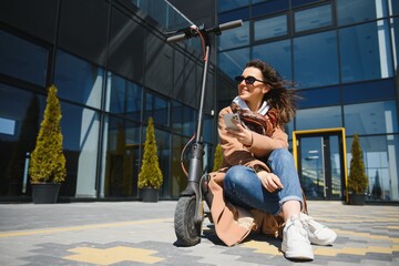 Beautiful young woman in sunglasses is sitting on her electro scooter near big glass building.