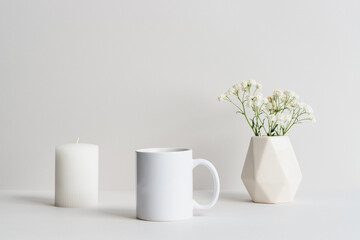 White mug mockup with beige vase, candle and gypsophila branch on a table